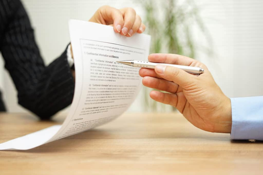 Close-up of two people exchanging a document and pen across a desk, preparing to sign an agreement or contract.