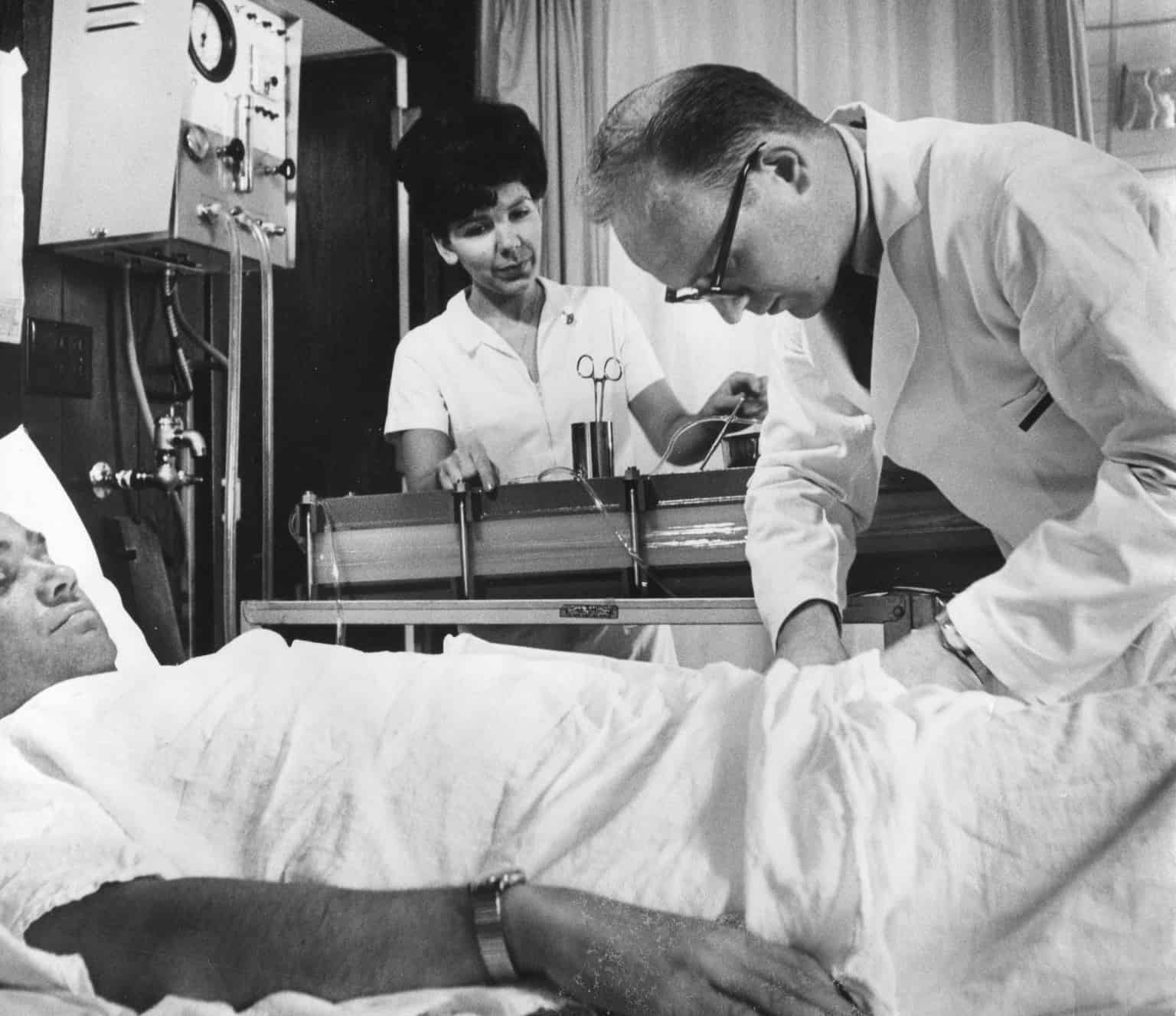 A black-and-white photo of a doctor examining a patient in a hospital bed while a nurse prepares medical instruments nearby. The scene captures mid-20th century hospital care with vintage medical equipment in the background.