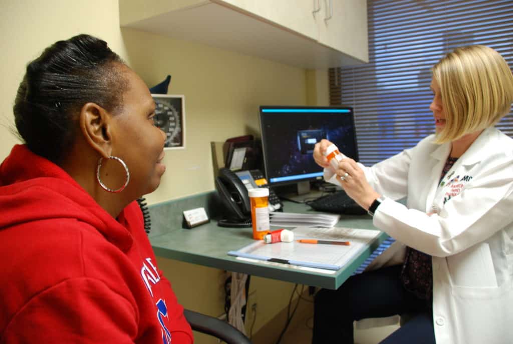 Pharmacist in a white coat consulting with a patient while organizing medication on a tray in a clinical office setting.
