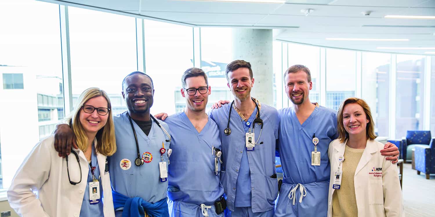 A diverse group of six smiling healthcare professionals in scrubs and white coats stand together with arms around each other in a bright hospital setting.