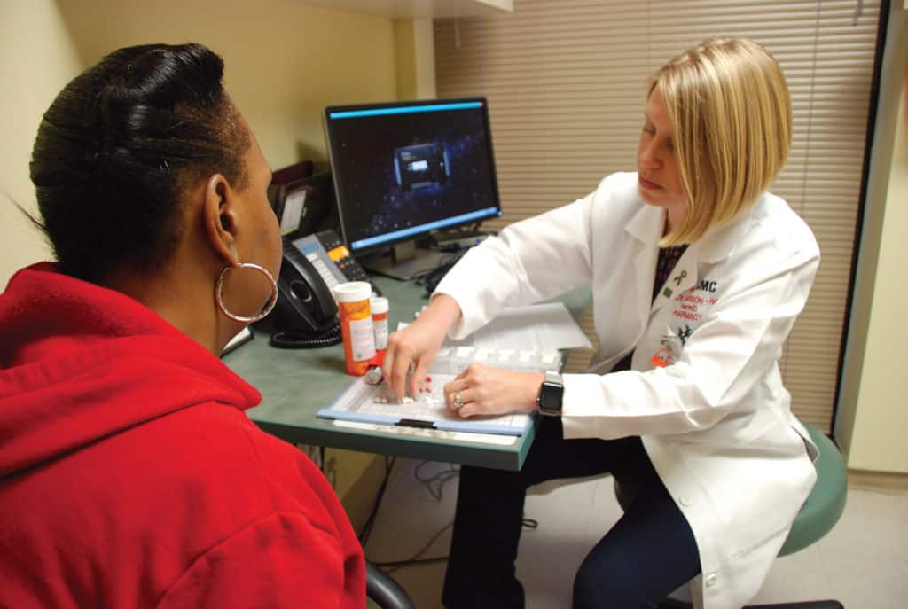 Pharmacist in a white coat consulting with a patient while organizing medication on a tray in a clinical office setting.