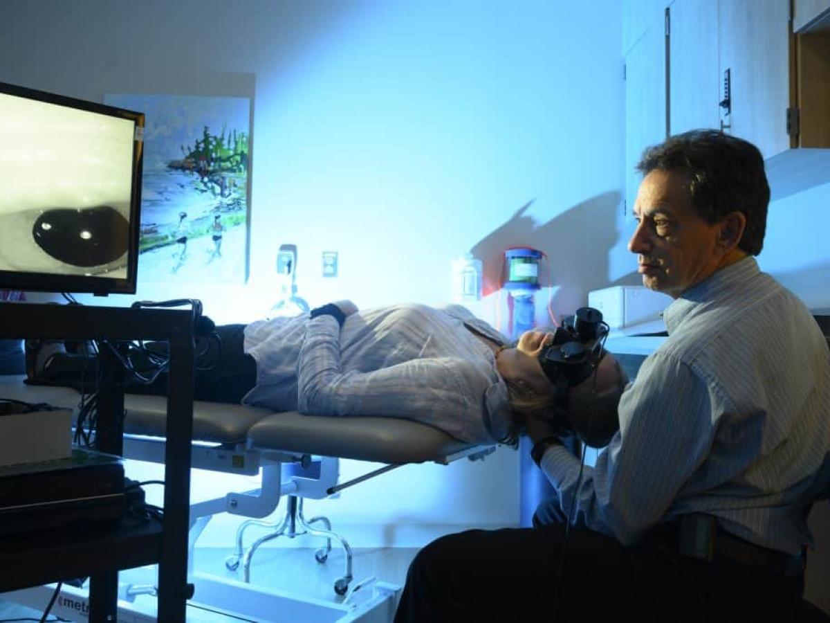 A doctor observing a screen as a patient is laying on a medical bed wearing a headset over their eyes in a dark room