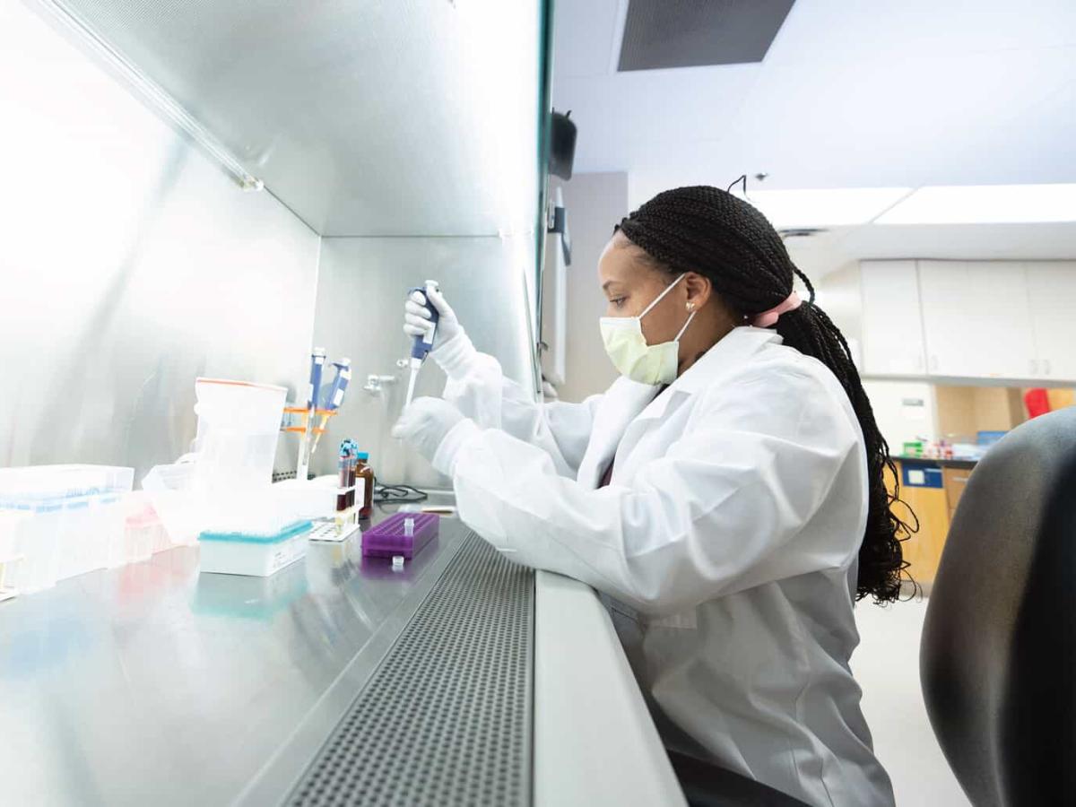 Lab technician wearing a mask and lab coat uses a pipette under a ventilation hood in a bright, sterile laboratory setting.