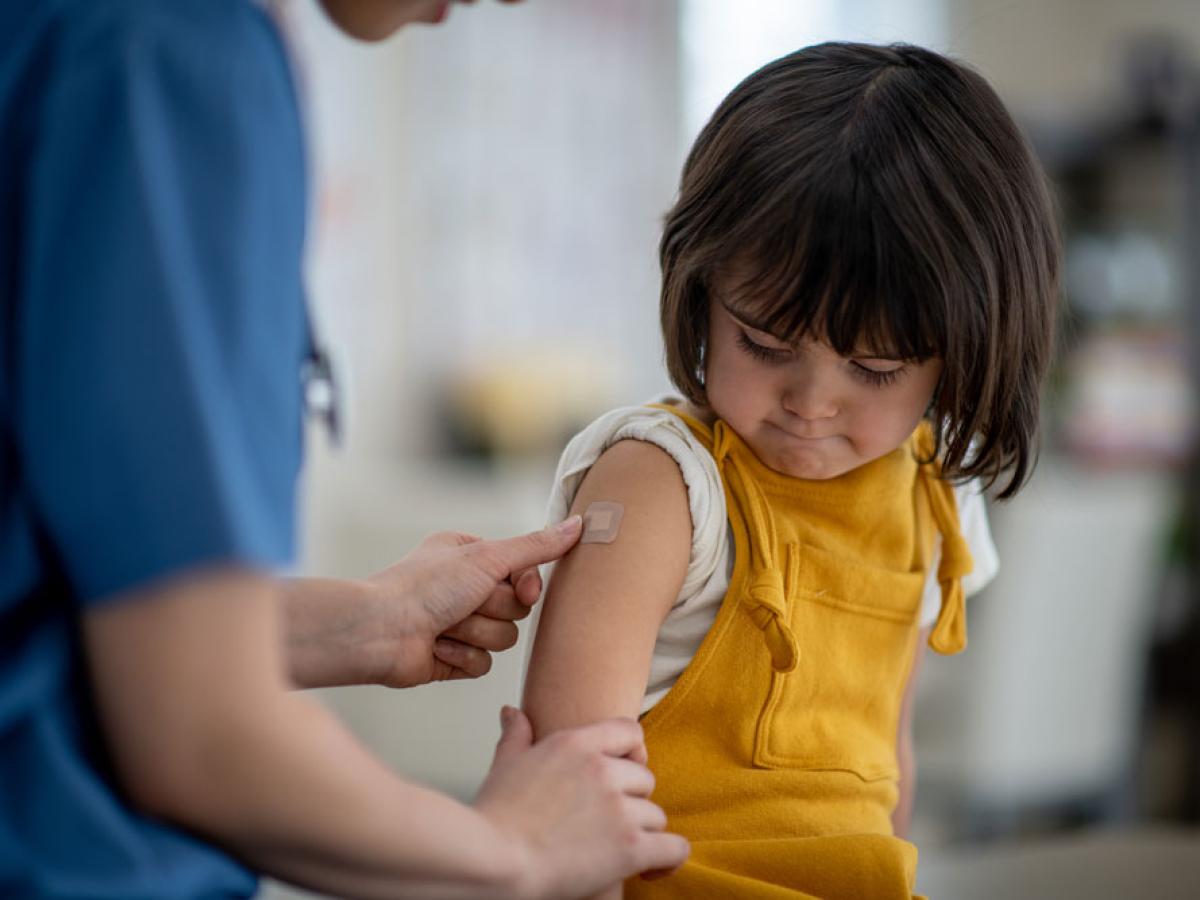 school-age girl in yellow coveralls getting vaccinated by nurse in blue scrubs