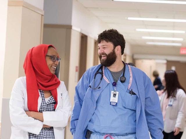  Two smiling healthcare providers, one in a red headscarf and white coat and the other in blue scrubs, walk and talk in a hospital hallway with other staff in the background.