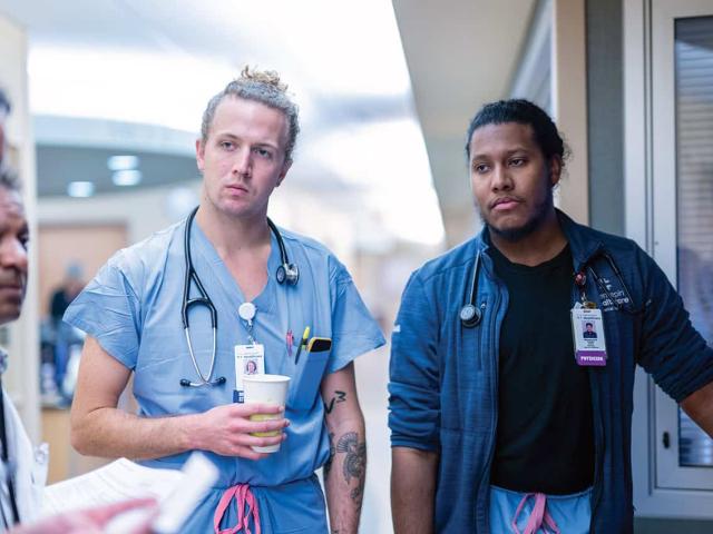 Medical residents and physicians in scrubs engaged in a team discussion during patient rounds in a hospital corridor.