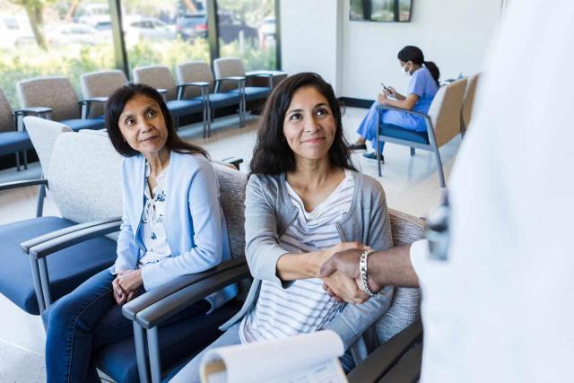 Two women, appearing to be a mother and daughter, are seated in a well-lit hospital waiting area. The mother, wearing a light blue cardigan, looks attentively to her right, while the daughter, dressed in a striped shirt and grey cardigan, smiles warmly and shakes hands with a medical professional whose arm is visible in the frame. In the background, a nurse in blue scrubs is seated, focused on a mobile device. The atmosphere is welcoming and calm, with large windows bringing in natural light.
