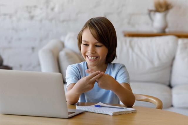 A young boy with light brown hair is sitting at a table in a cozy, well-lit living room. He is smiling while practicing sign language in front of a laptop. A notepad and pen are on the table in front of him, suggesting he is engaged in an online learning session. The background features a white sofa and soft decor, creating a calm and welcoming environment.