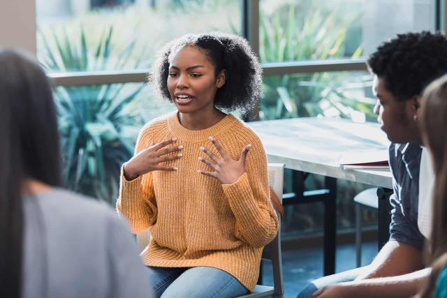 A young woman wearing a mustard-colored sweater is speaking expressively during a group therapy session. She is seated in a bright, open room with large windows and greenery visible outside. Other participants, slightly out of focus, are listening attentively, creating an atmosphere of support and openness.