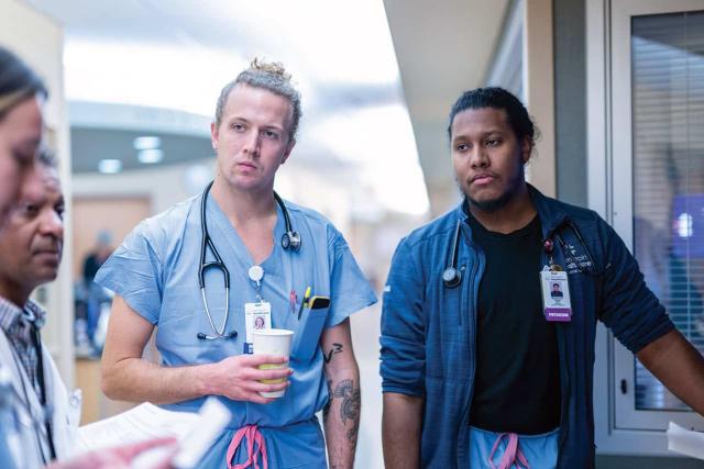 Medical residents and physicians in scrubs engaged in a team discussion during patient rounds in a hospital corridor.