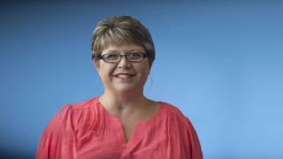 woman with glasses and pink blouse smiling against a blue background
