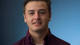 Male in a burgundy shirt smiling against a blue background
