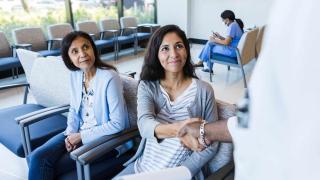 Two women, appearing to be a mother and daughter, are seated in a well-lit hospital waiting area. The mother, wearing a light blue cardigan, looks attentively to her right, while the daughter, dressed in a striped shirt and grey cardigan, smiles warmly and shakes hands with a medical professional whose arm is visible in the frame. In the background, a nurse in blue scrubs is seated, focused on a mobile device. The atmosphere is welcoming and calm, with large windows bringing in natural light.