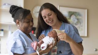 Two smiling nurses in blue scrubs are gently caring for a newborn baby. One nurse is holding the baby, while the other adjusts the infant’s hat. They are in a warm, softly lit birth center or hospital room.