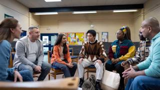 A diverse group of people sits in a circle of chairs in a community room, engaged in a group discussion or support meeting.