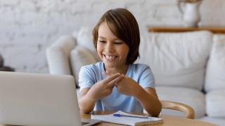 A young boy with light brown hair is sitting at a table in a cozy, well-lit living room. He is smiling while practicing sign language in front of a laptop. A notepad and pen are on the table in front of him, suggesting he is engaged in an online learning session. The background features a white sofa and soft decor, creating a calm and welcoming environment.