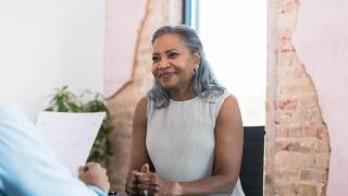 An older woman with gray hair sits at a desk, smiling and engaged in conversation with someone across from her who is holding a document. 