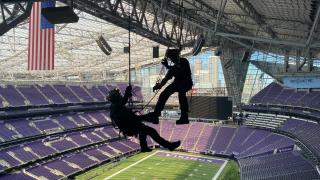 Two EMS staff members suspended by harnesses perform a high-angle rescue training exercise inside U.S. Bank Stadium. The stadium’s purple seats, American flag, and Minneapolis skyline are visible in the background.