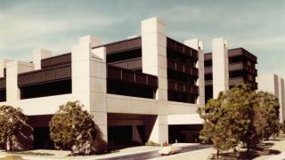 Architectural rendering from 1976 of the Hennepin County Medical Center, showing a modernist concrete and glass hospital building with surrounding trees, driveways, and parked vehicles.