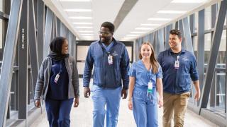 A diverse group of four healthcare professionals walk together down a bright hospital corridor, smiling and engaged in conversation. They wear Hennepin Healthcare uniforms and ID badges, conveying a sense of teamwork and camaraderie.
