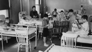 Black-and-white photo from the 1930s showing children in hospital beds reading books and magazines during library time in a pediatric ward. A nurse or librarian is assisting, with a book cart labeled "Hospital Service" in the center.