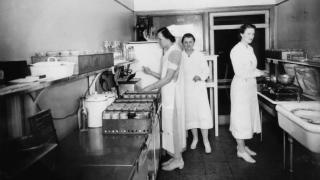 Black-and-white photo from 1938 showing three women in white uniforms working in a hospital milk formula laboratory. They prepare bottles and use vintage kitchen equipment in a clean, tiled workspace.