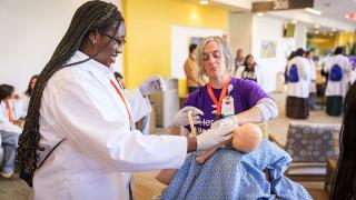 A young woman in a white lab coat and gloves smiles as she practices a medical procedure on an infant mannequin. She is guided by an instructor wearing a purple “Hennepin Healthcare” shirt and gloves, who is holding a medical tool. The training takes place in a bright, active space with other participants in the background, all wearing lab coats, suggesting a hands-on educational healthcare event.
