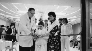 A black-and-white photo of a ribbon-cutting ceremony in a pediatric playroom. A child, assisted by a doctor and a woman, cuts the ribbon while staff and attendees look on and smile in the background.