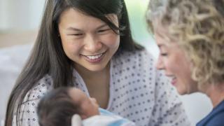 A young mother in a hospital gown smiles warmly while looking at her newborn baby, who is swaddled in a hospital blanket. Beside her, a nurse or healthcare professional with curly blonde hair also smiles while admiring the baby. 