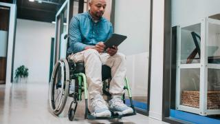 Man in wheelchair using tablet in modern accessible healthcare facility, demonstrating digital patient engagement and inclusive medical technology solutions.