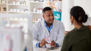 Pharmacist in a white coat smiling and explaining medication instructions to a patient across the counter in a pharmacy setting.
