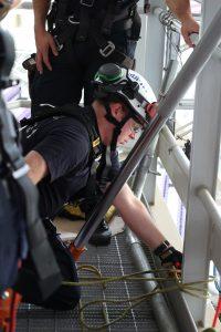A rescue worker in a helmet and safety harness kneels on a grated platform, securing ropes during a training exercise. Another team member stands nearby, both wearing black uniforms and tactical gear.