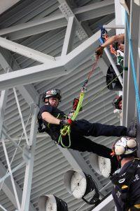 A rescue worker in safety gear and helmet is suspended by ropes during a high-angle training exercise inside a stadium, with structural beams and other team members visible in the background.
