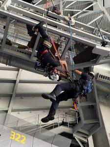 Two emergency responders in harnesses perform a suspended rope rescue training drill beneath a metal catwalk inside a stadium. One rescuer appears to assist the other near section 322.