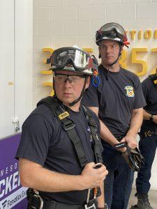 Two emergency response team members in safety helmets and harnesses stand indoors, appearing focused during a training session. One holds a radio while the other looks on attentively.