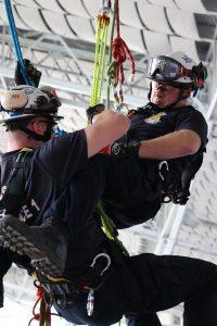 Two emergency rescue personnel perform a rope training exercise while suspended in the air. Both are wearing helmets, harnesses, and safety gear inside a large indoor stadium structure.