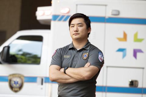 A Hennepin EMS student stands confidently with arms crossed in front of an ambulance bearing the Hennepin Healthcare logo, wearing a uniform that identifies him as a paramedic student.