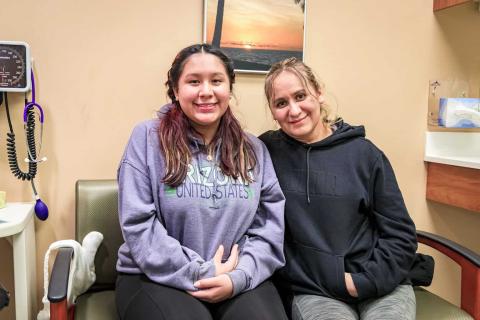A smiling teenage girl and an adult woman sit side by side in a clinic exam room, showing a sense of comfort and connection during a healthcare visit.