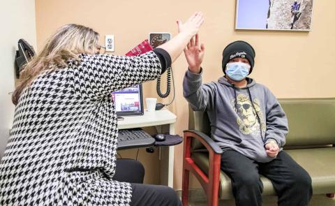 A healthcare staff member and a young patient wearing a mask share a high-five in a clinic exam room, celebrating a positive interaction.