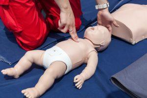 Person demonstrating infant CPR on a training mannequin, using two fingers to apply compressions to the center of the infant’s chest.