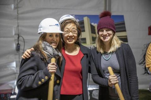 Three women, two wearing hard hats and one in a knit hat, smile and pose with wooden groundbreaking shovels at a construction event for the Redleaf Center.