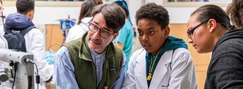 A young student in a white lab coat observes a medical monitor alongside an adult mentor and another learner, engaging in a hands-on healthcare education experience in a clinical setting.