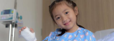 Young girl in a hospital gown with cartoon animals smiles while sitting in a hospital bed, holding up a bandaged hand with an IV line, with medical equipment visible in the background.