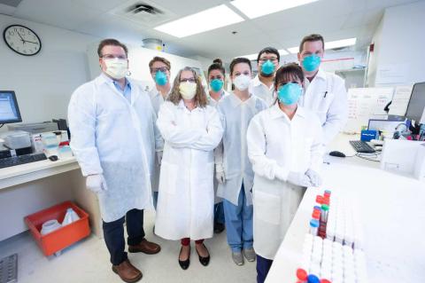 Group of laboratory staff in protective masks and lab coats standing together in a clinical lab setting, with medical equipment and test tubes visible in the foreground.
