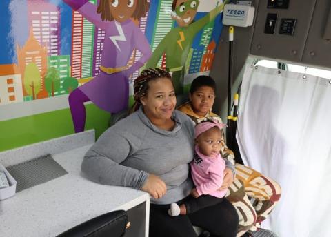 A mother sits with her two children inside the Pediatric Mobile Clinic, with colorful superhero-themed artwork on the wall behind them.