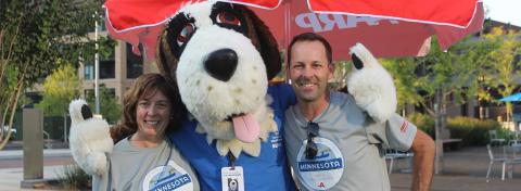 Two Hennepin Healthcare staff members smile and pose with Bernie the St. Bernard mascot during a community outreach event, standing under a red umbrella.