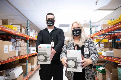 Two supply chain employees in a medical storage room holding boxes of 3M N95 respirators, wearing branded face masks that say "Supply Chain... We Have You Covered."