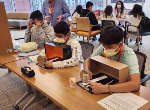 Students wearing masks participate in a hands-on medical learning activity using simulated surgical tools and shadow boxes in a classroom setting.