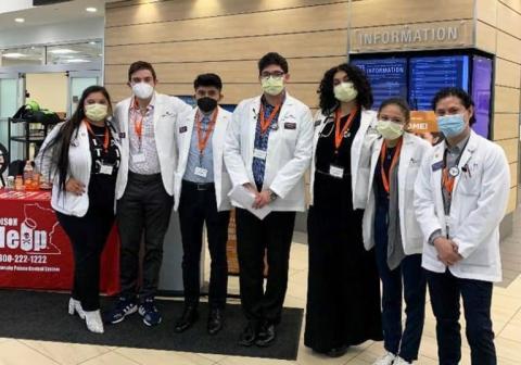 A group of students in lab coats and face masks pose together in a hospital or clinic lobby, standing in front of an information desk during a health education event.