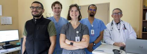 A diverse group of five healthcare professionals, including doctors and medical staff, smile while standing confidently in a clinical setting with computers and medical equipment visible in the background.
